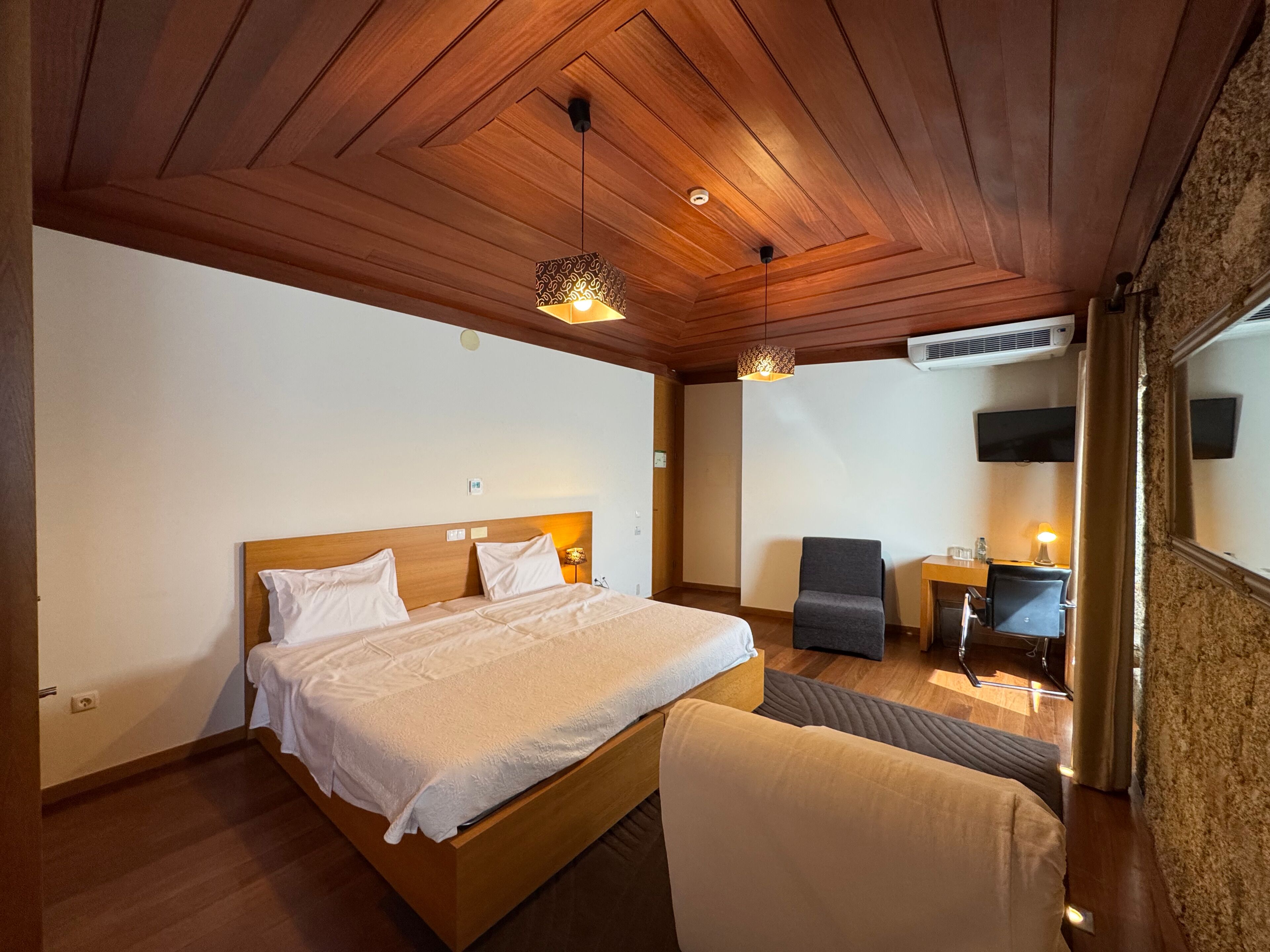 Un dormitorio con techo de madera, una cama con ropa de cama blanca, un escritorio con silla y un televisor montado en la pared.