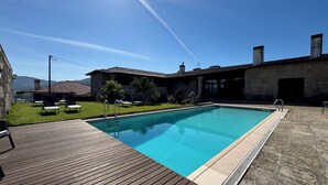 A rectangular swimming pool with a wooden deck, surrounded by a stone building with a tiled roof, and a clear blue sky. - Hotel Alves (Braga)