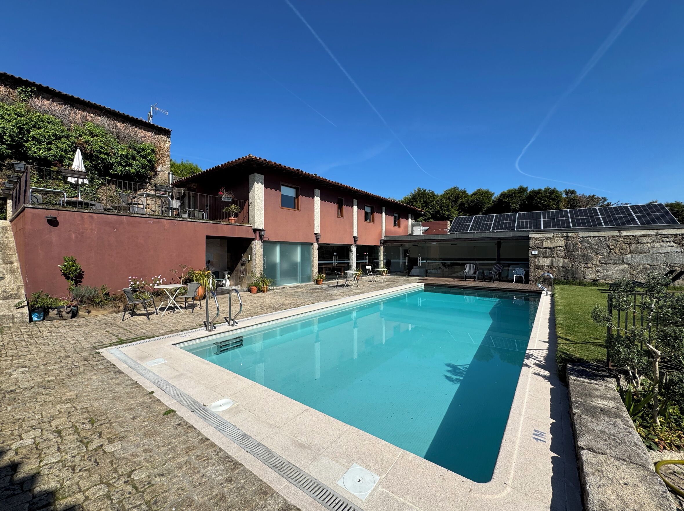 A swimming pool with a stone deck, a red building with a balcony, and solar panels on the roof.