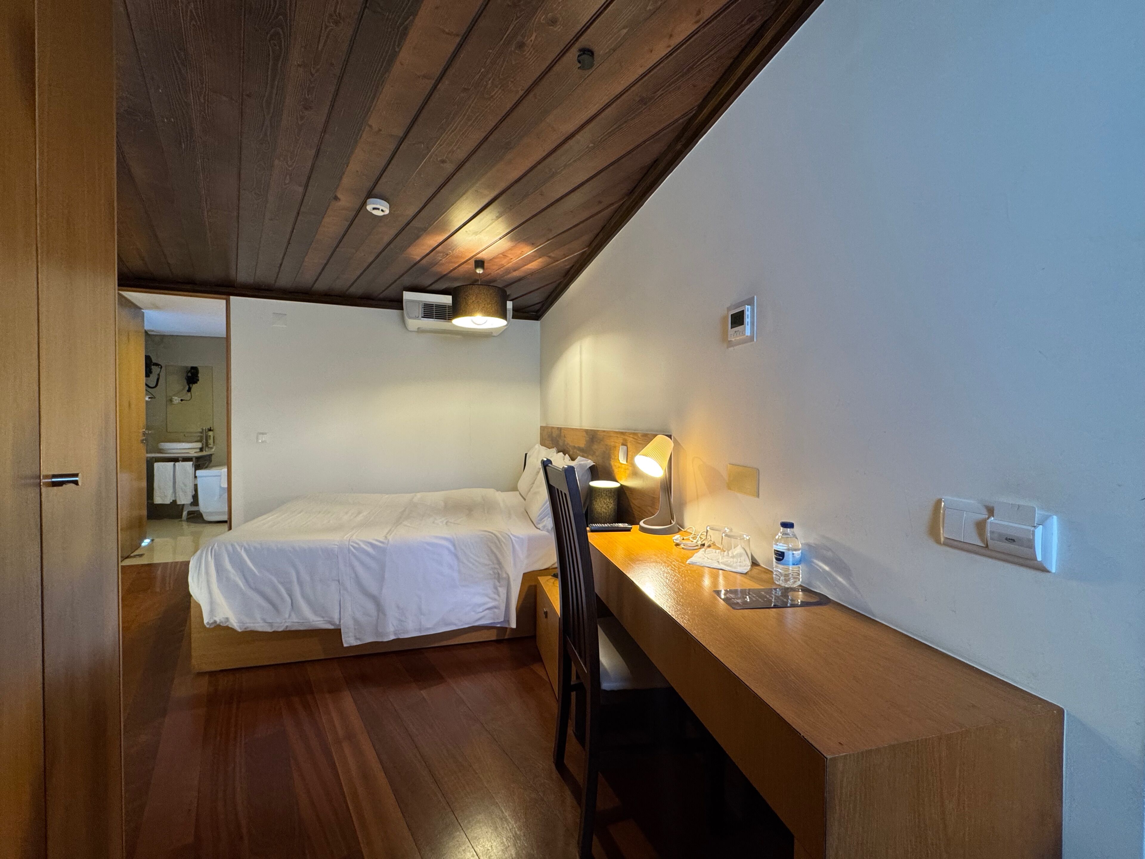 Una habitación con una cama, un escritorio con una lámpara y un espejo. Tiene techo de madera y aire acondicionado instalado en la pared.
