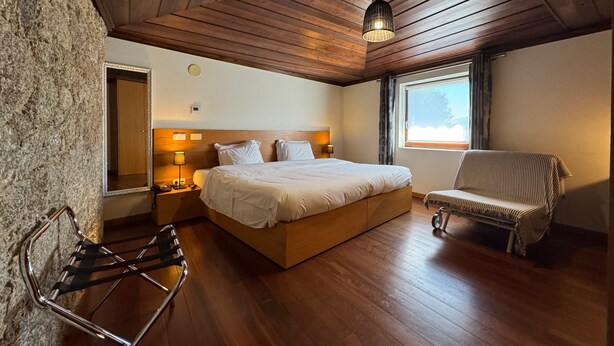 A bedroom with a wooden ceiling, a bed with white linens, a chair, a nightstand, and a window with curtains. - Hotel Alves (Braga)