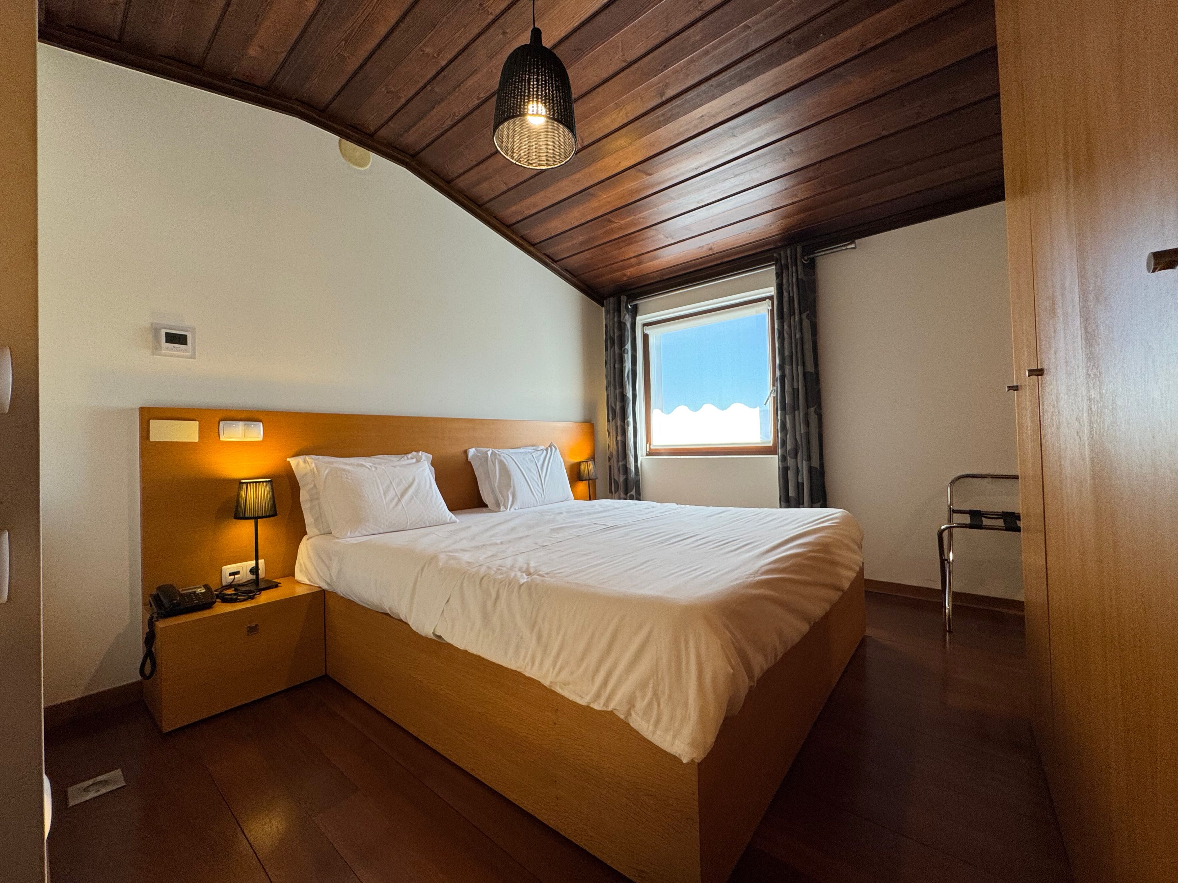 Ein Schlafzimmer mit Holzbalkendecke, einem Bett mit weißer Bettwäsche, einem Nachttisch mit Lampe, einem Thermostat und einem Fenster mit Vorhängen.