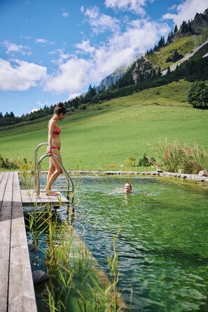 Eine Holzterrasse führt zu einem klaren Pool, auf dem eine Person steht und eine andere im Wasser.