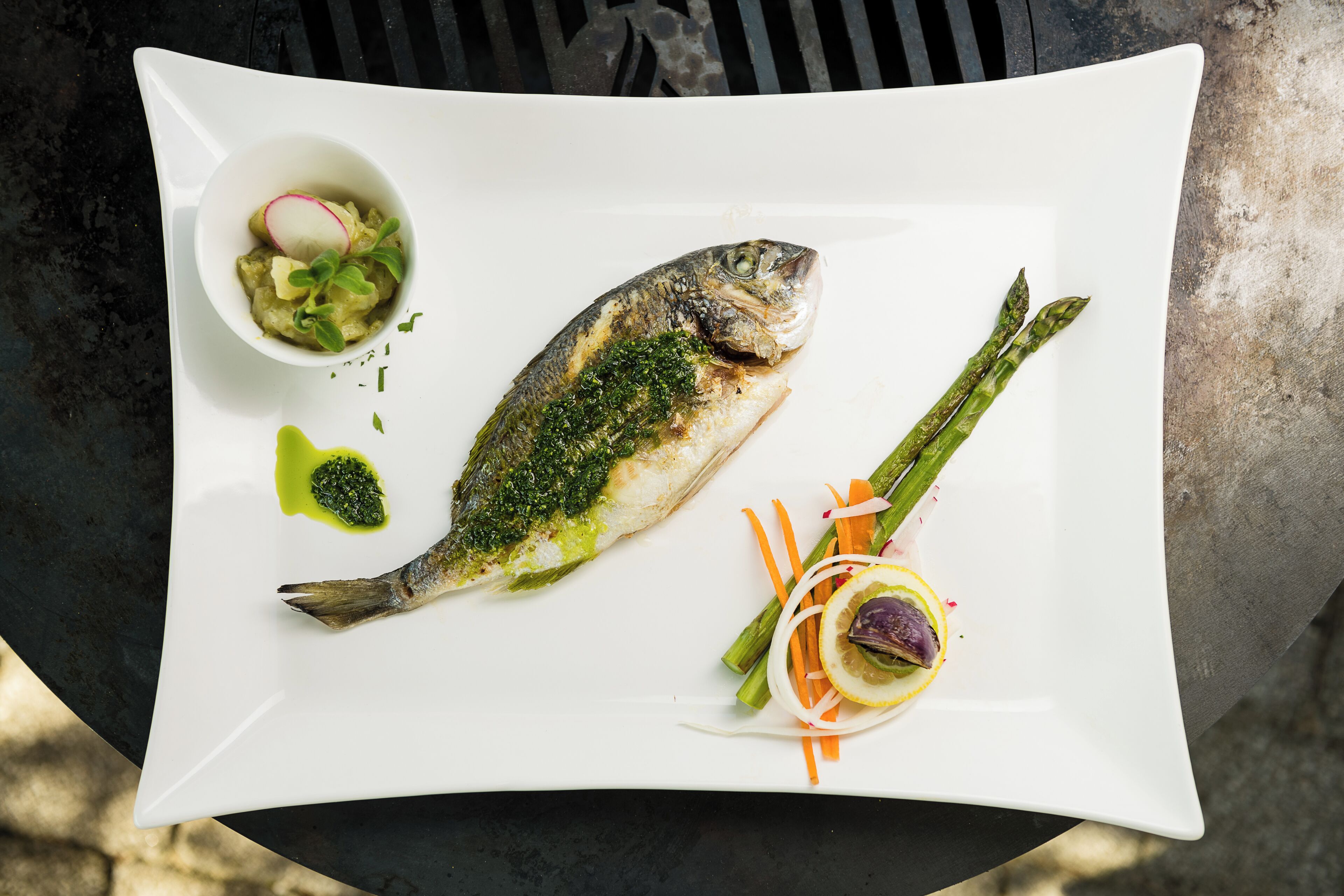 Un plato blanco cuadrado con un pescado a la parrilla, espárragos y un cuenco pequeño de ensalada.