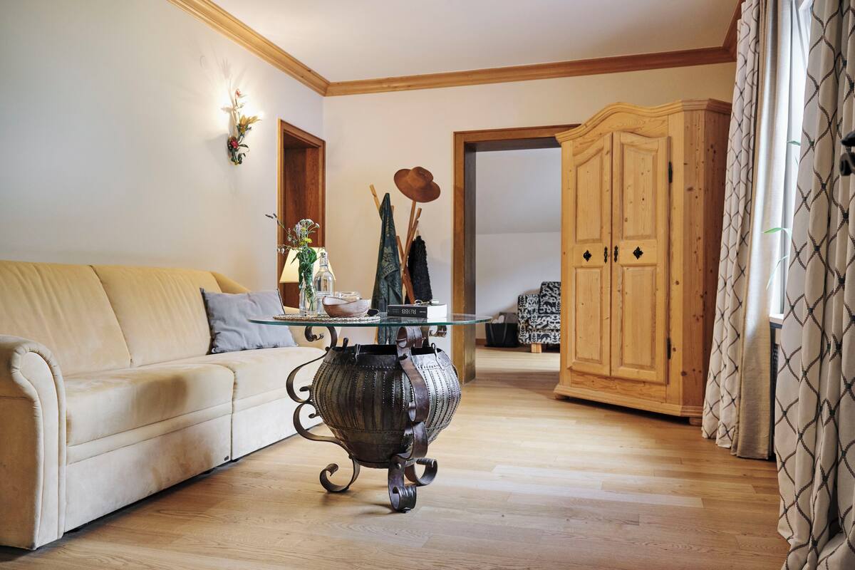 Een woonkamer met een beige bank, een salontafel met glazen blad, een decoratieve metalen pot, houten kasten en een gordijn met patroon.