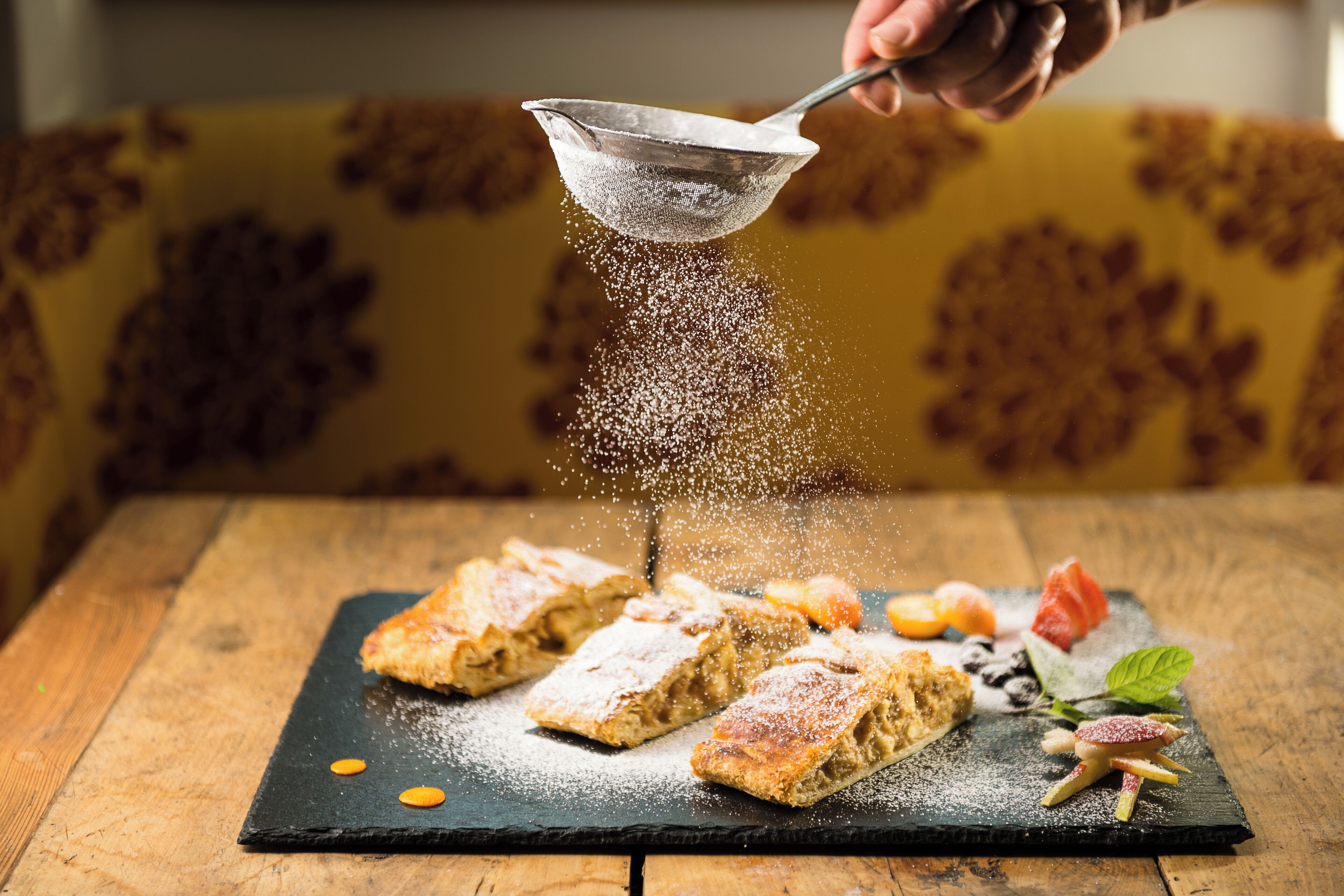 A dessert with powdered sugar being sprinkled, accompanied by fruit and mint leaves on a slate serving board.
