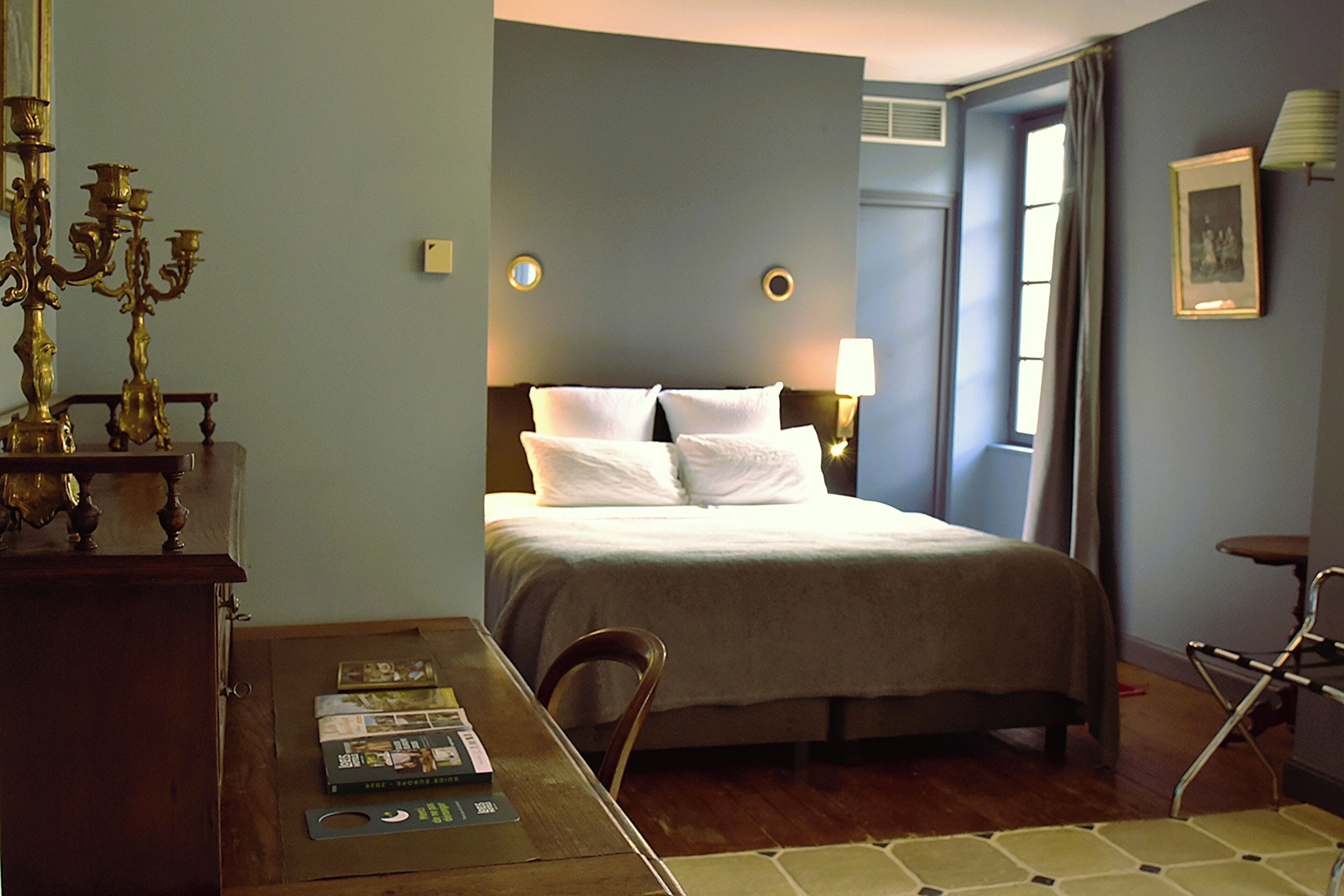 A hotel room with a bed, a wooden table with magazines, a lamp, and a candelabra.