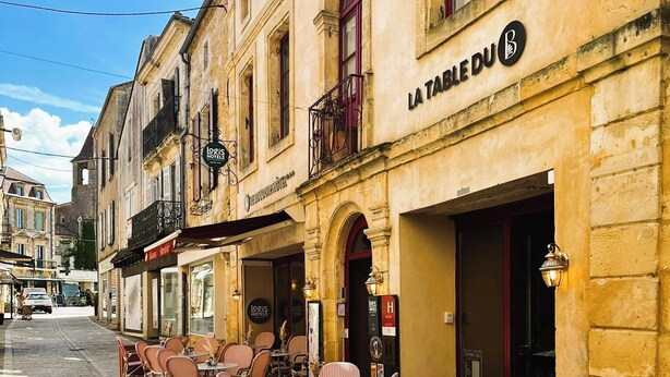 Une rue avec des tables en terrasse, une enseigne indiquant « RESTAURANT, SALON DE THÉ, BAR – COCKTAILS, GLACES DU PÉRIGORD », et un bâtiment portant l’inscription « LOGIS HOTELS ».