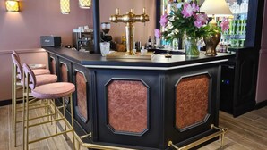 Un espace bar comprenant un comptoir, des tabourets de bar, une machine à café, un moulin à café, une cafetière, un verre, un vase avec des fleurs et une étagère avec des bouteilles.
