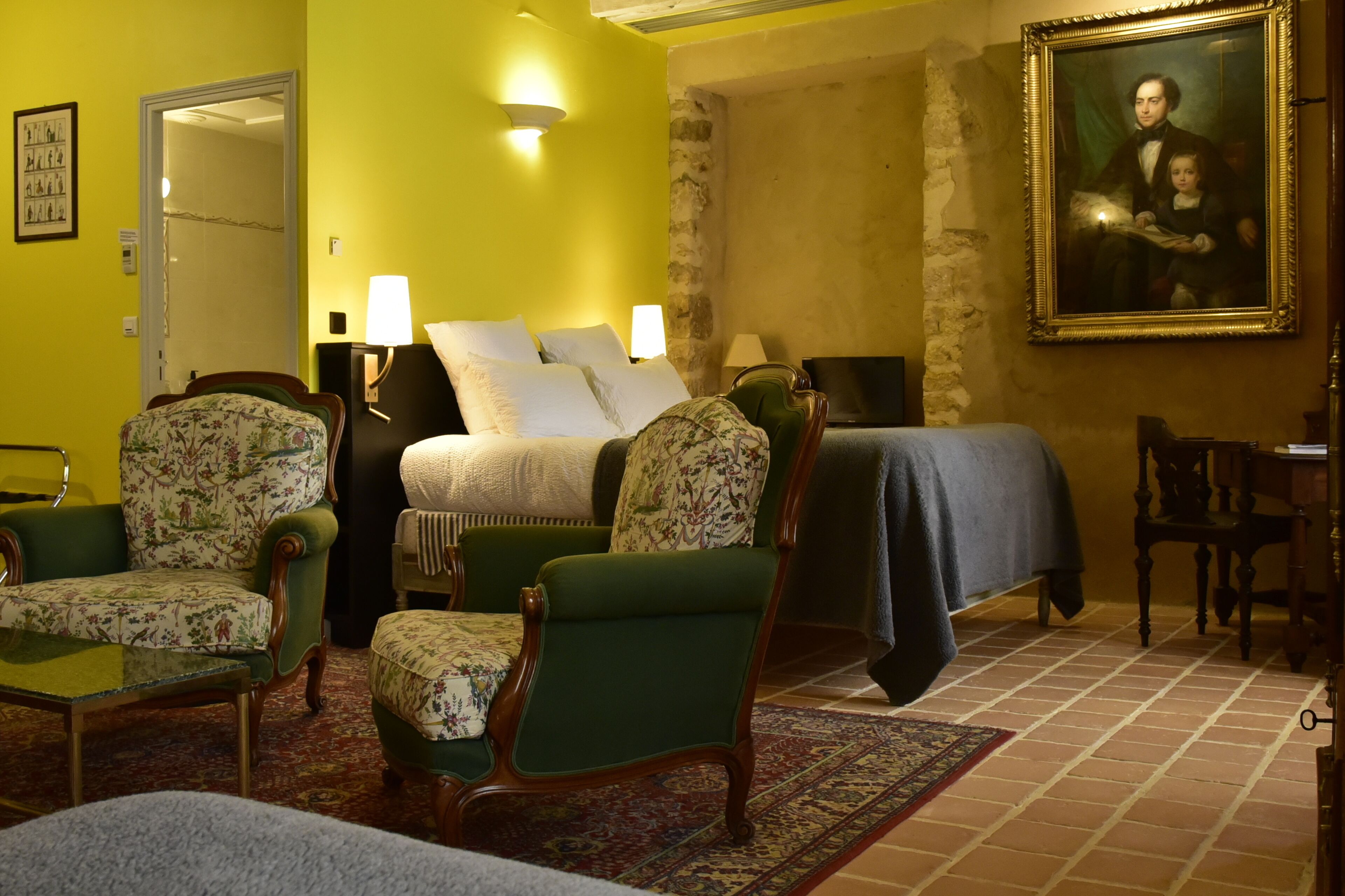 Una habitación con una cama, dos sillas, una mesa, una lámpara, un cuadro y una puerta.