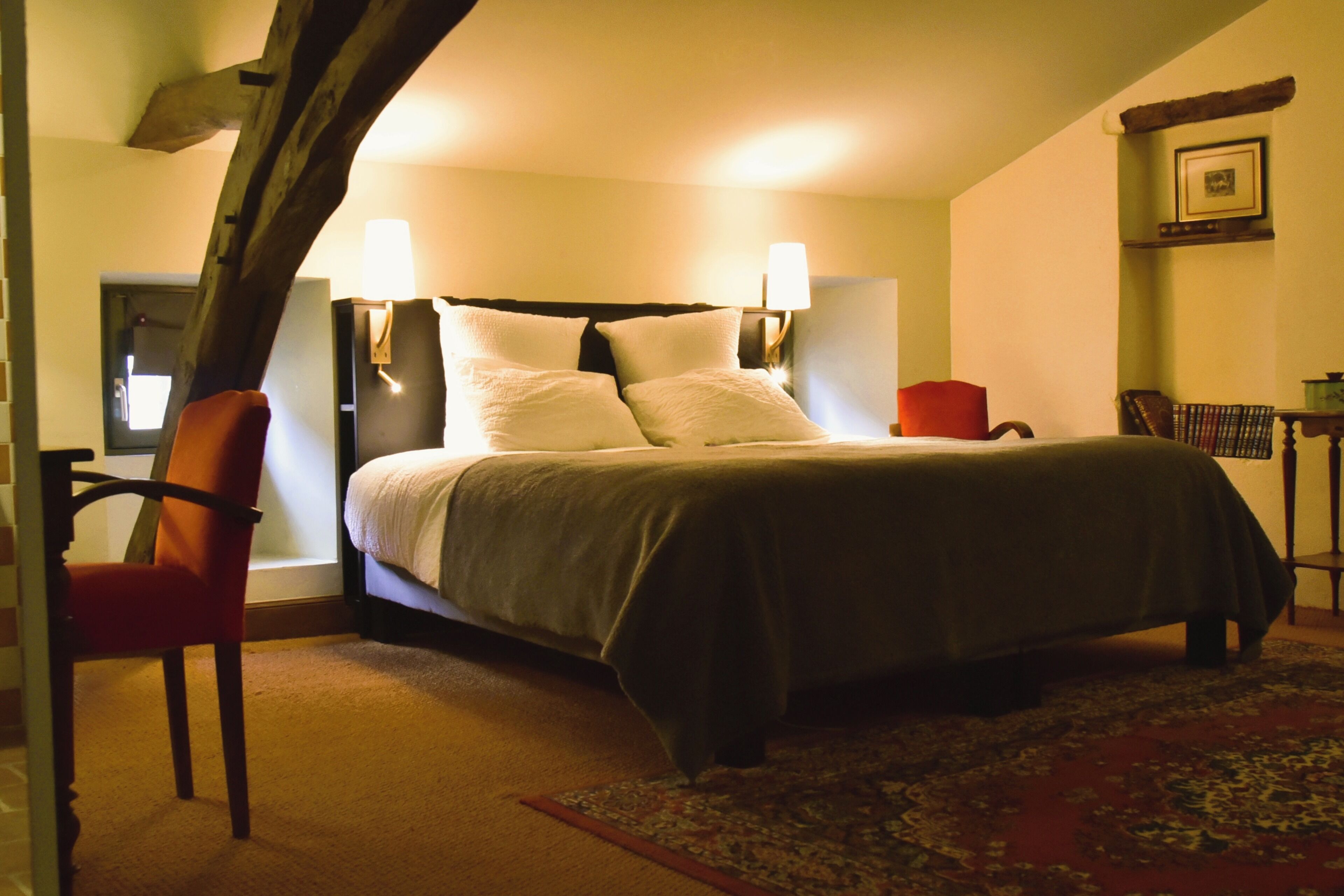 A bed with a headboard, two bedside lamps, a chair, a small table, and a rug in a room with a sloped ceiling and a picture on the wall.