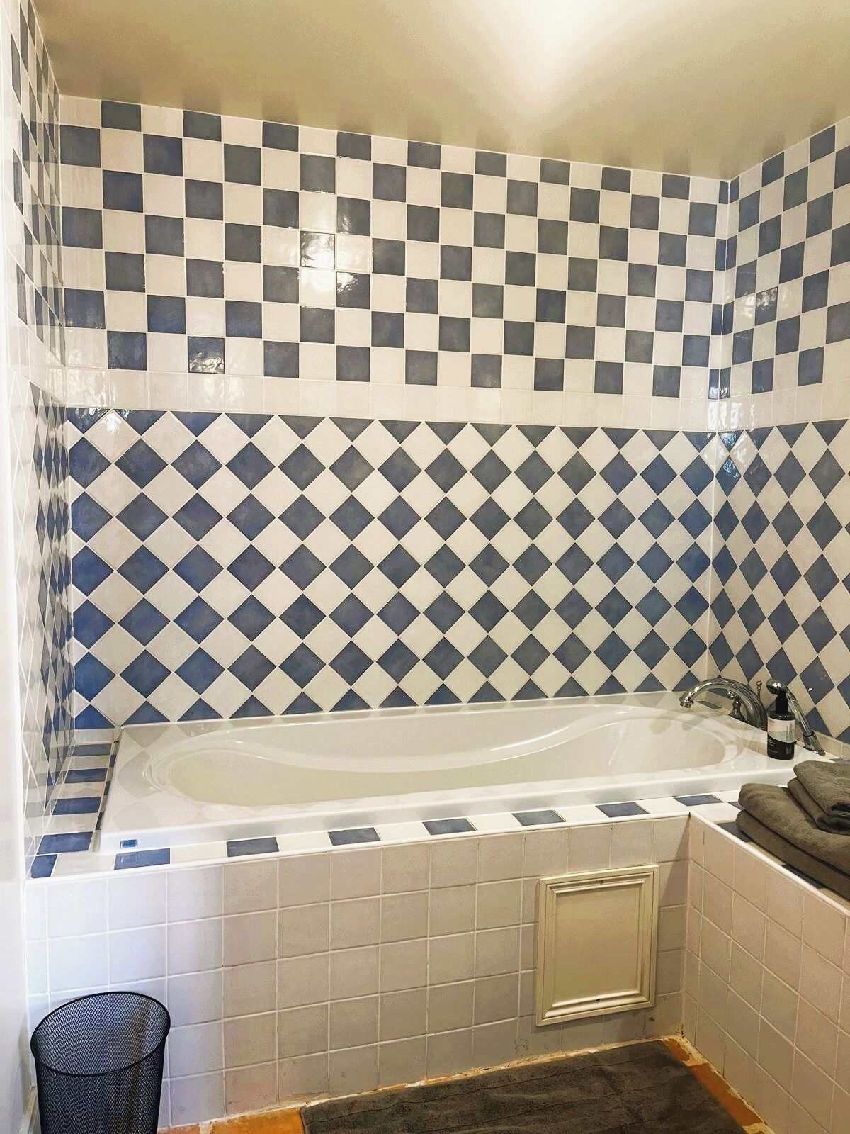 A bathroom with a checkered tile wall, a bathtub, and a small trash bin.