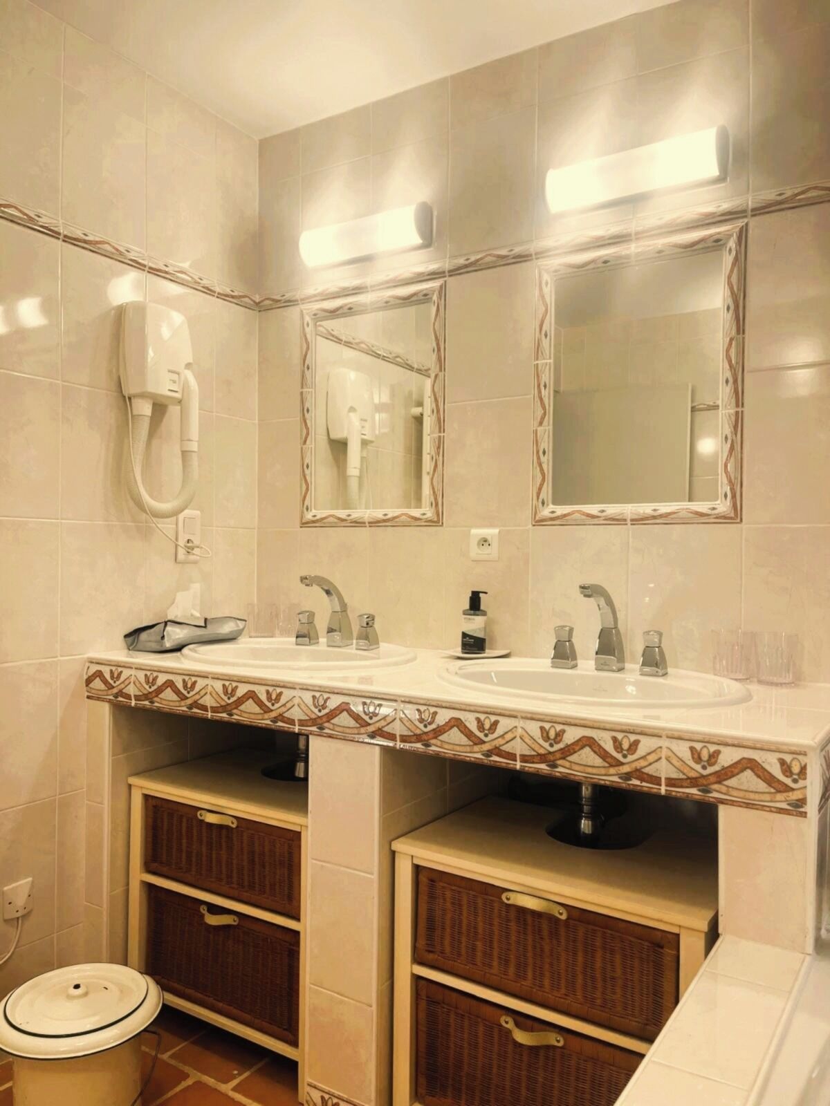 A bathroom with a double sink, two mirrors, a hairdryer, and a trash bin.