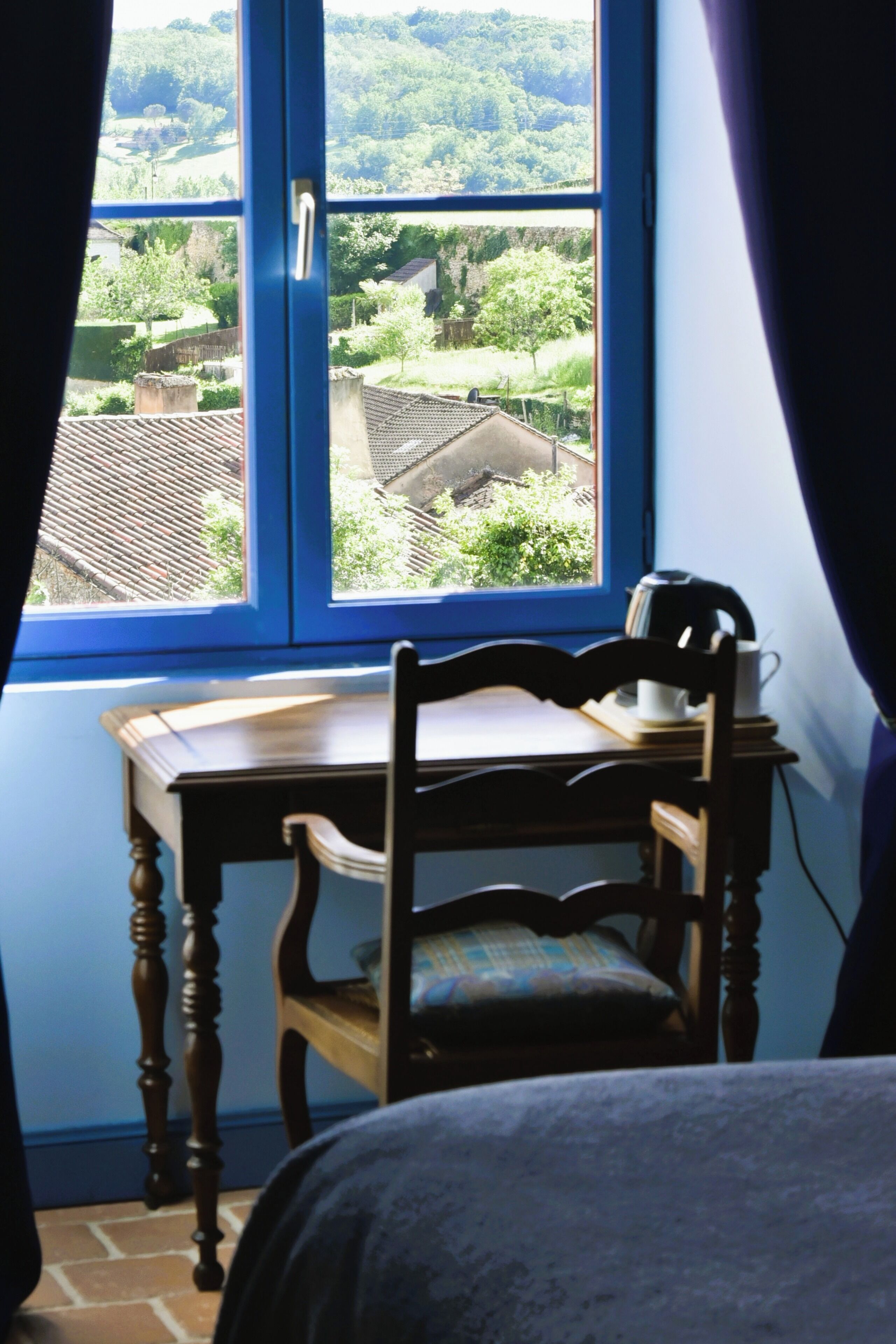 Een kamer met een raam dat uitkijkt op een groen landschap, een houten stoel en een tafel met een waterkoker.