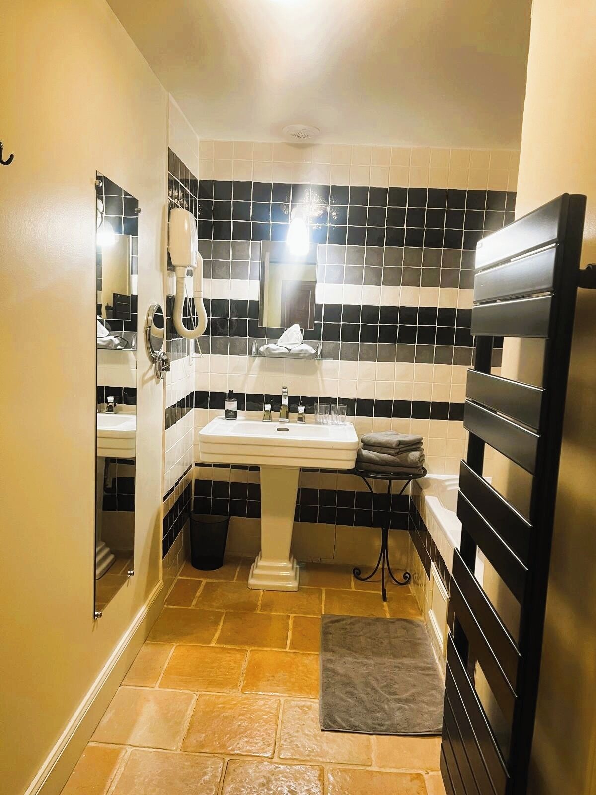 Un baño con un lavamanos blanco pedestal, paredes con azulejos blanco y negro, un perchero negro y un pequeño cubo de basura.