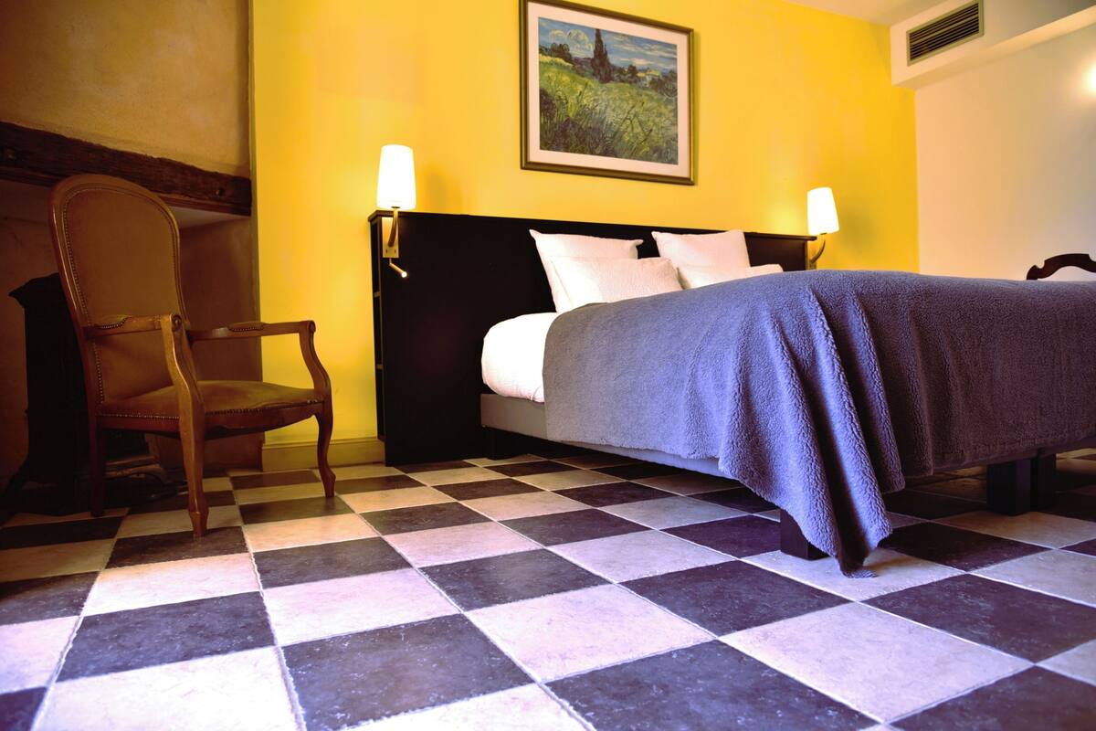 Een hotelkamer met een geruit vloerkleed, een bed met een donkere hoofdeinde, twee bedlampjes en een stoel.