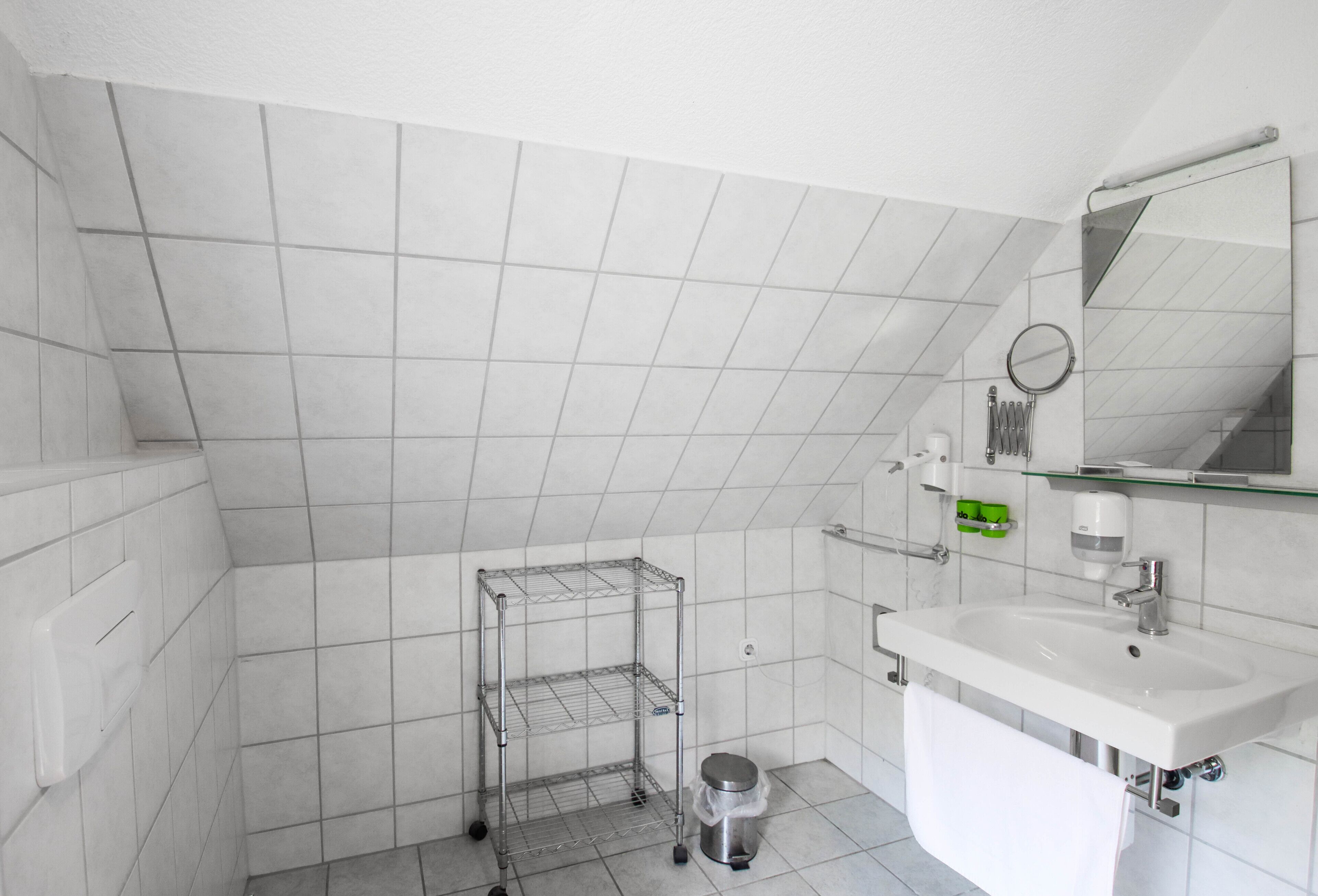 Une petite salle de bain aux murs recouverts de carreaux blancs, avec un seul lavabo, un miroir et une étagère en métal.