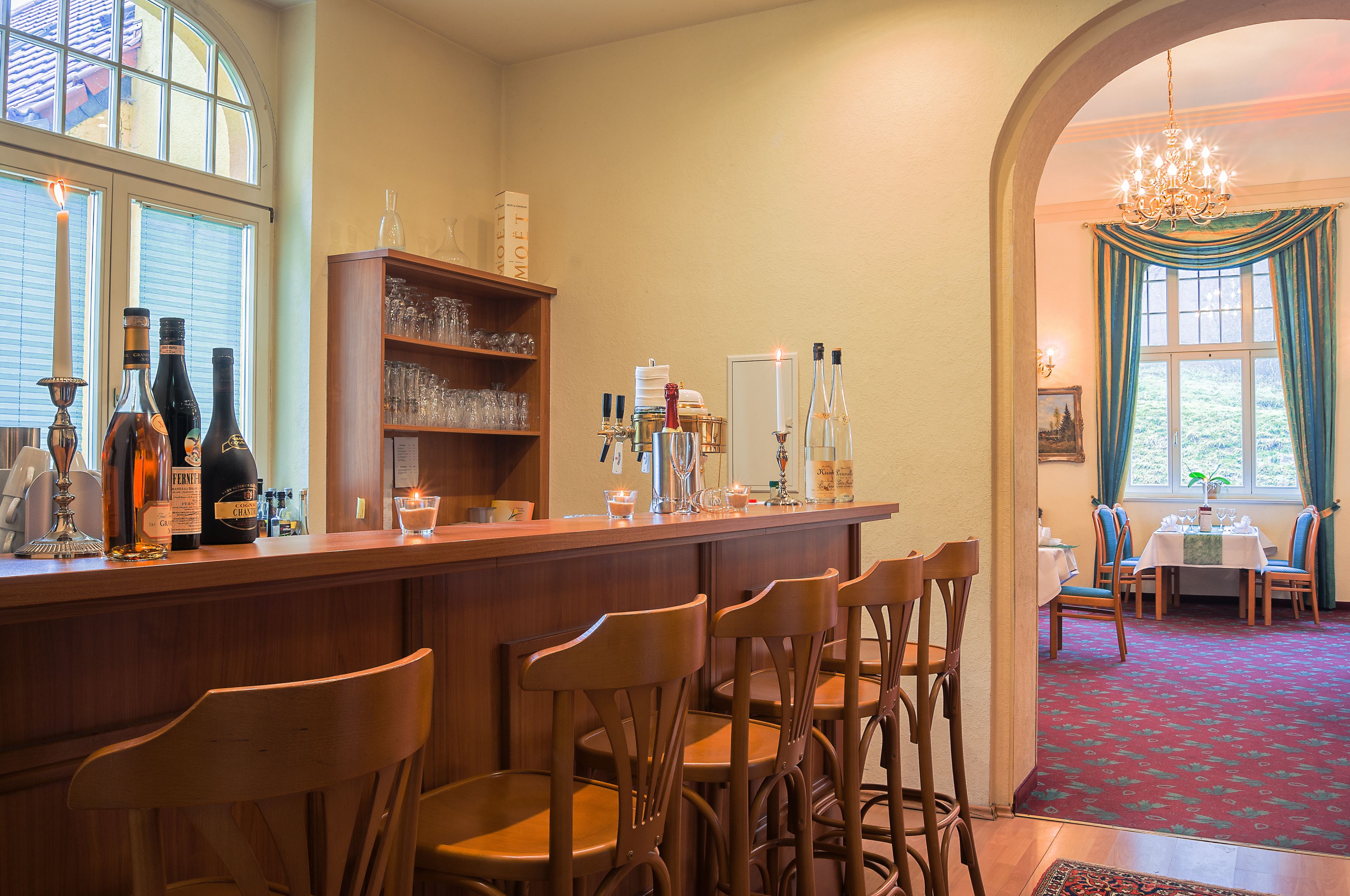 A bar area with wooden chairs, shelves with glasses and bottles, and a view of a dining area with a table and chairs.
