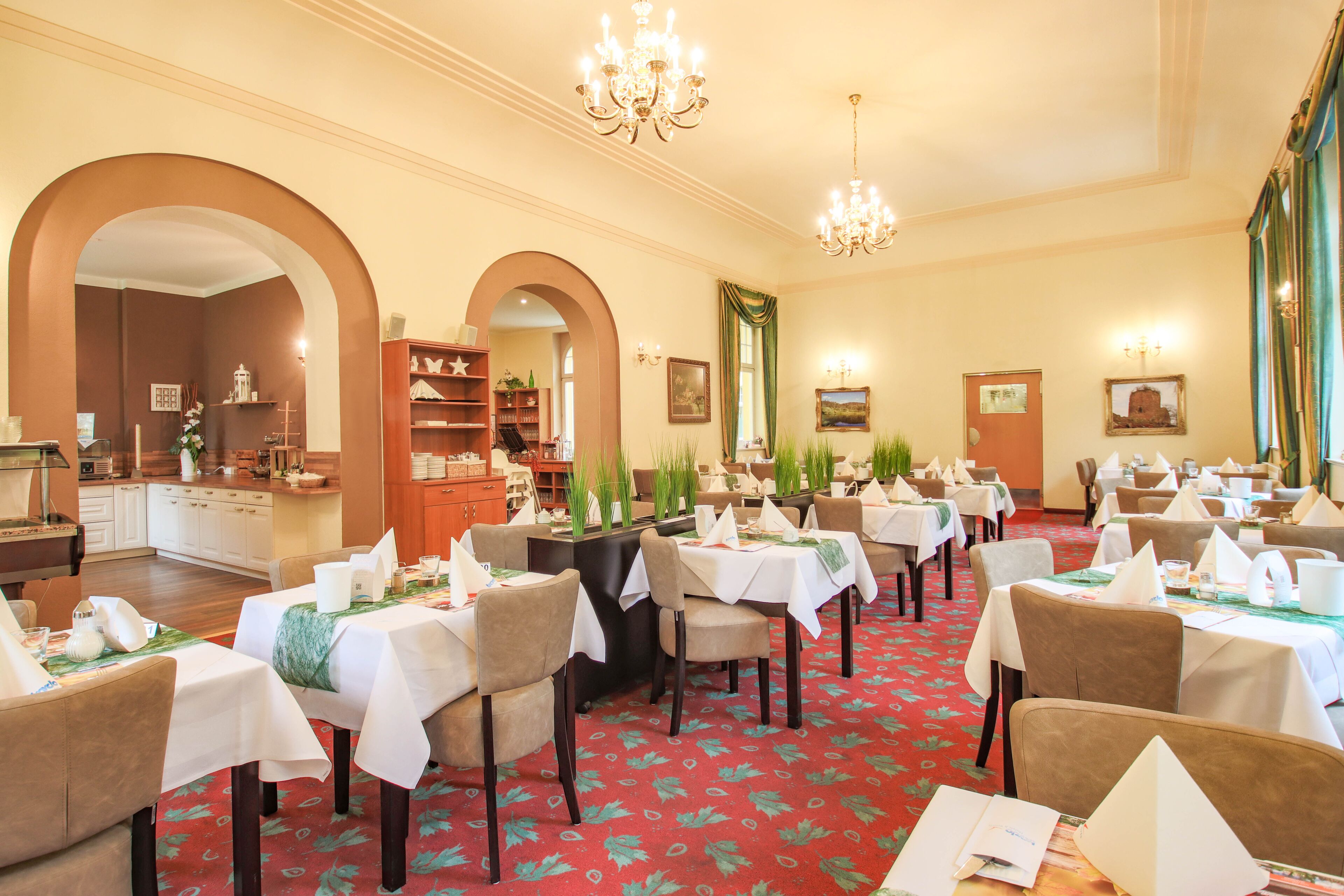 A dining area with tables set for service, a kitchen in the background, and a chandelier.