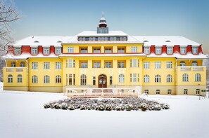 A large, yellow building with multiple windows and a central entrance, surrounded by snow. - Ferien Hotel Südharz (Ellrich)