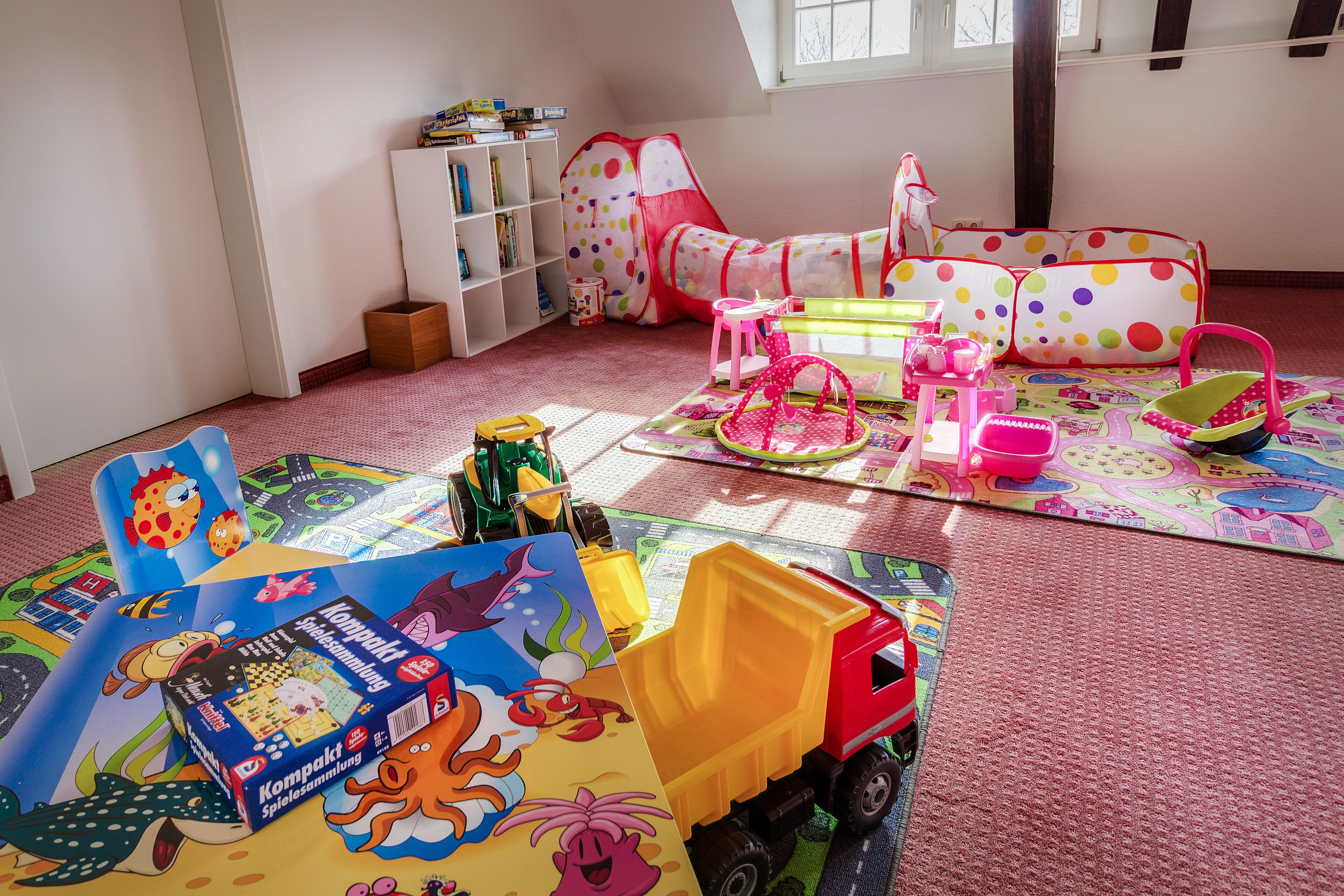 Uma sala de brinquedos com tapetes coloridos, uma estante de livros, uma tenda e um caminhão de brinquedo.