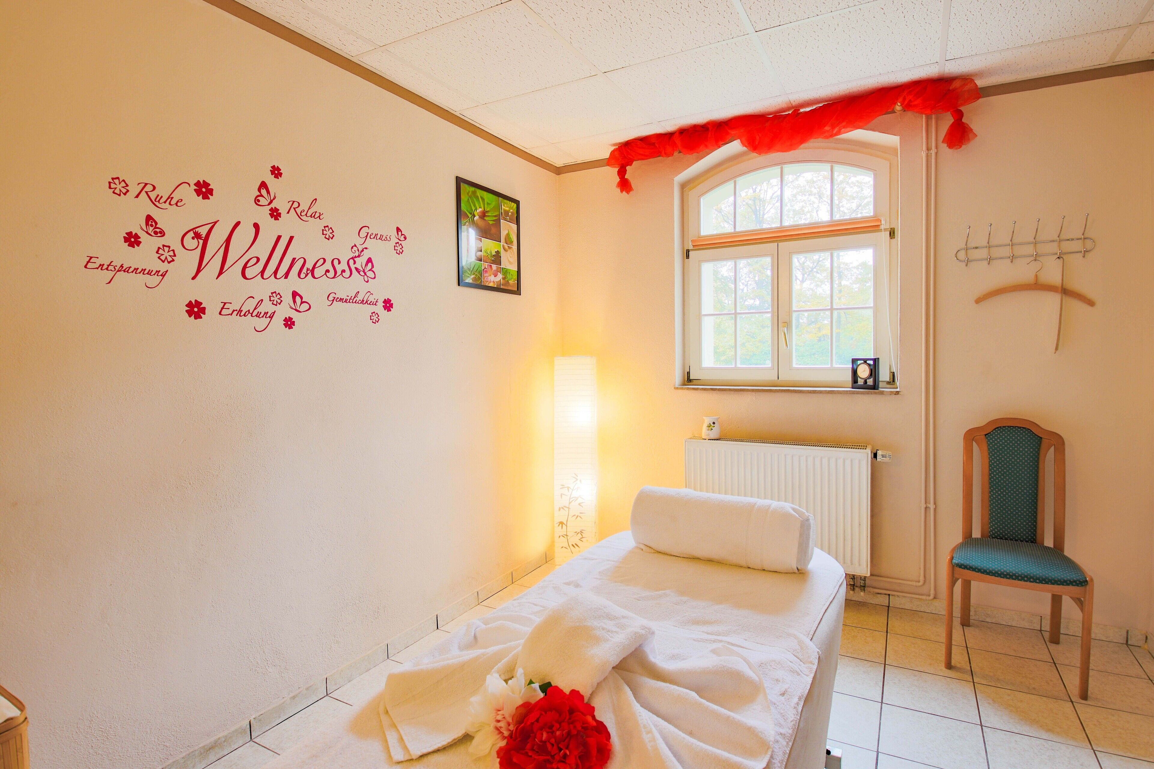 A bedroom with a bed, a chair, a window, a radiator, and a wall with wellness-related decorations.