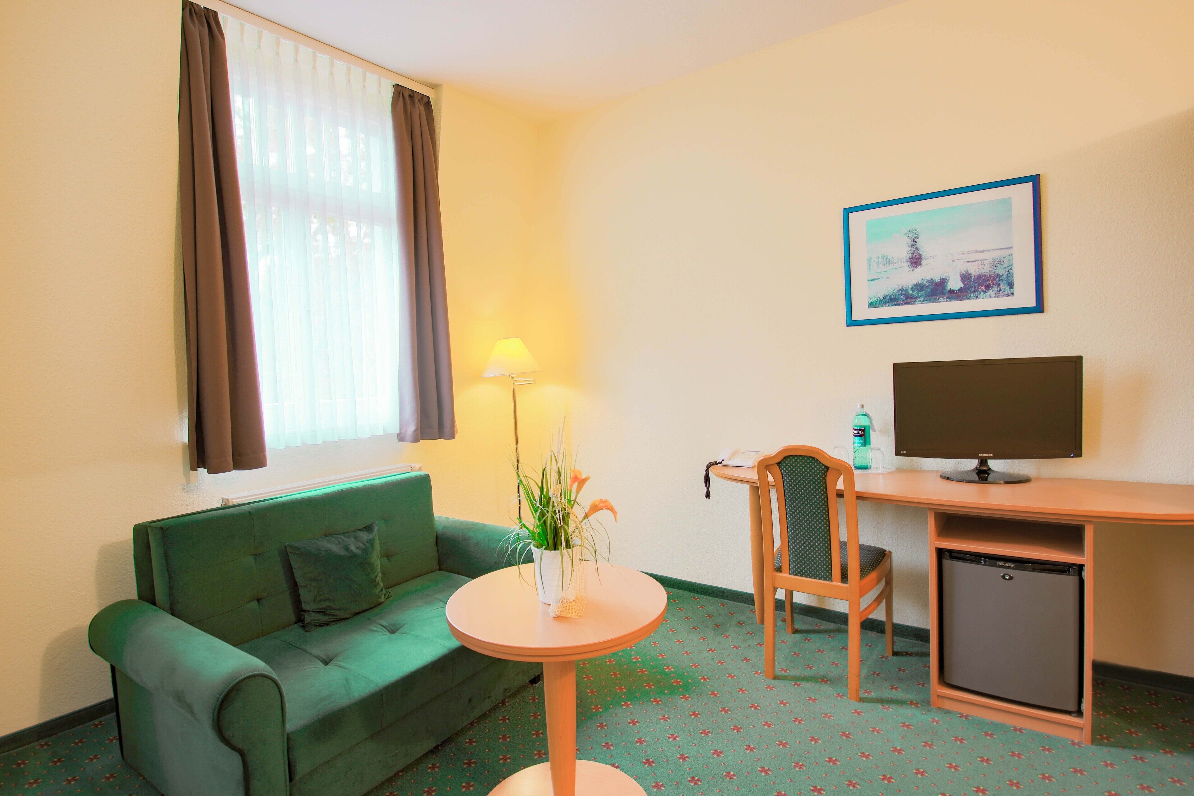 Een kamer met een groene bank, een klein rond tafeltje met een plant, een bureau met een computer en een kleine koelkast.