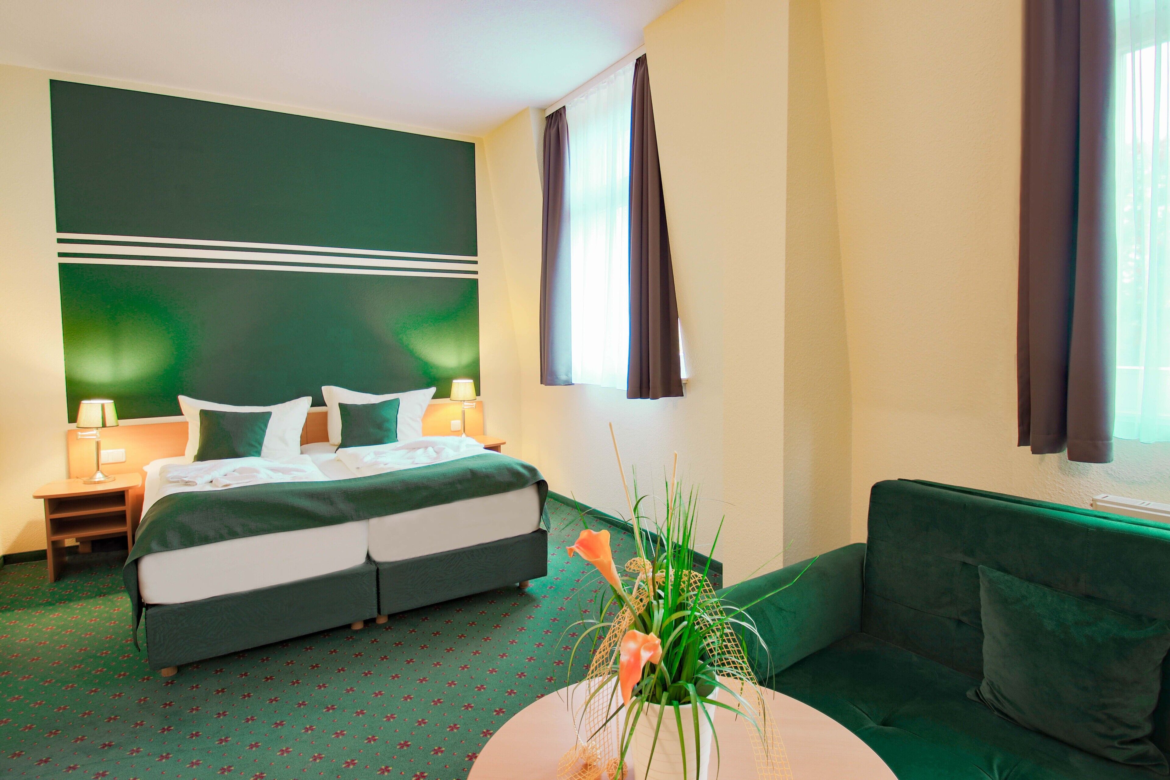 Een hotelkamer met een bed, een groene bank, een ronde tafel met een vaas, en een nachtkastje met een lamp.