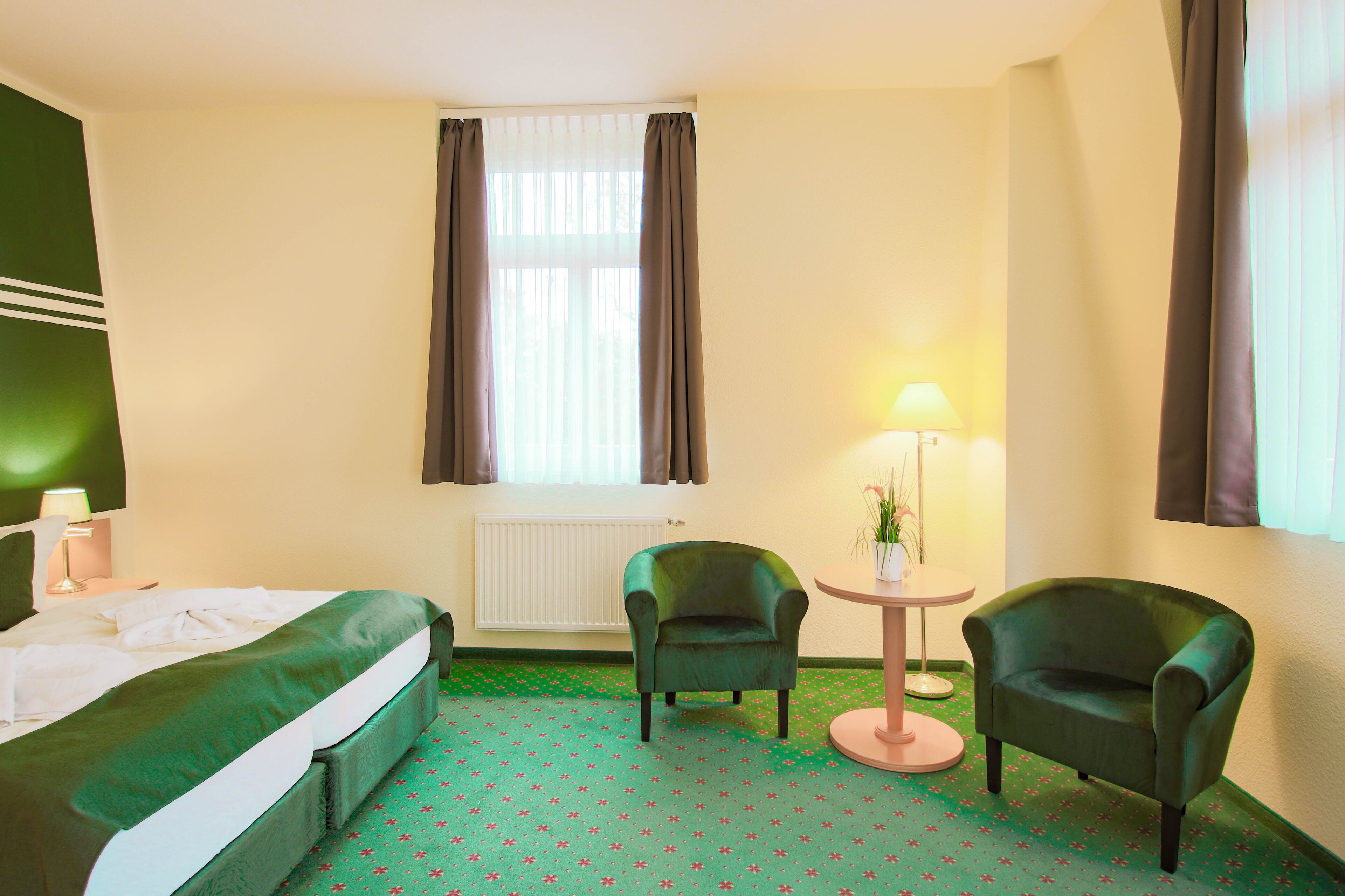 A hotel room with a bed, two chairs, a small table with a lamp, and a window with curtains.
