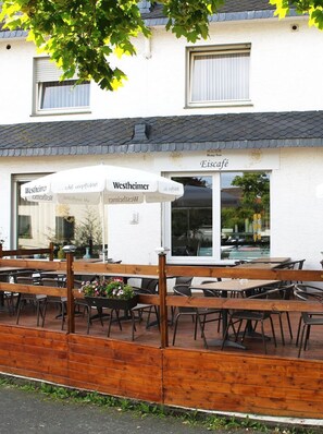 An outdoor seating area with wooden benches and tables, a white building with a sign reading "Eiscafé," and a large umbrella providing shade. - Hotel Haarener Hof (Bad Wuennenberg)