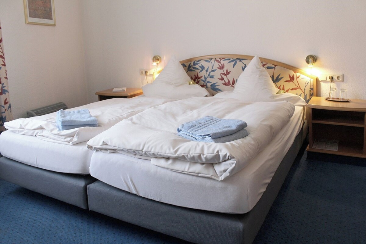 A neatly made bed with white linens and two folded towels, a wooden headboard with a floral pattern, bedside tables with lamps, and a blue carpet.