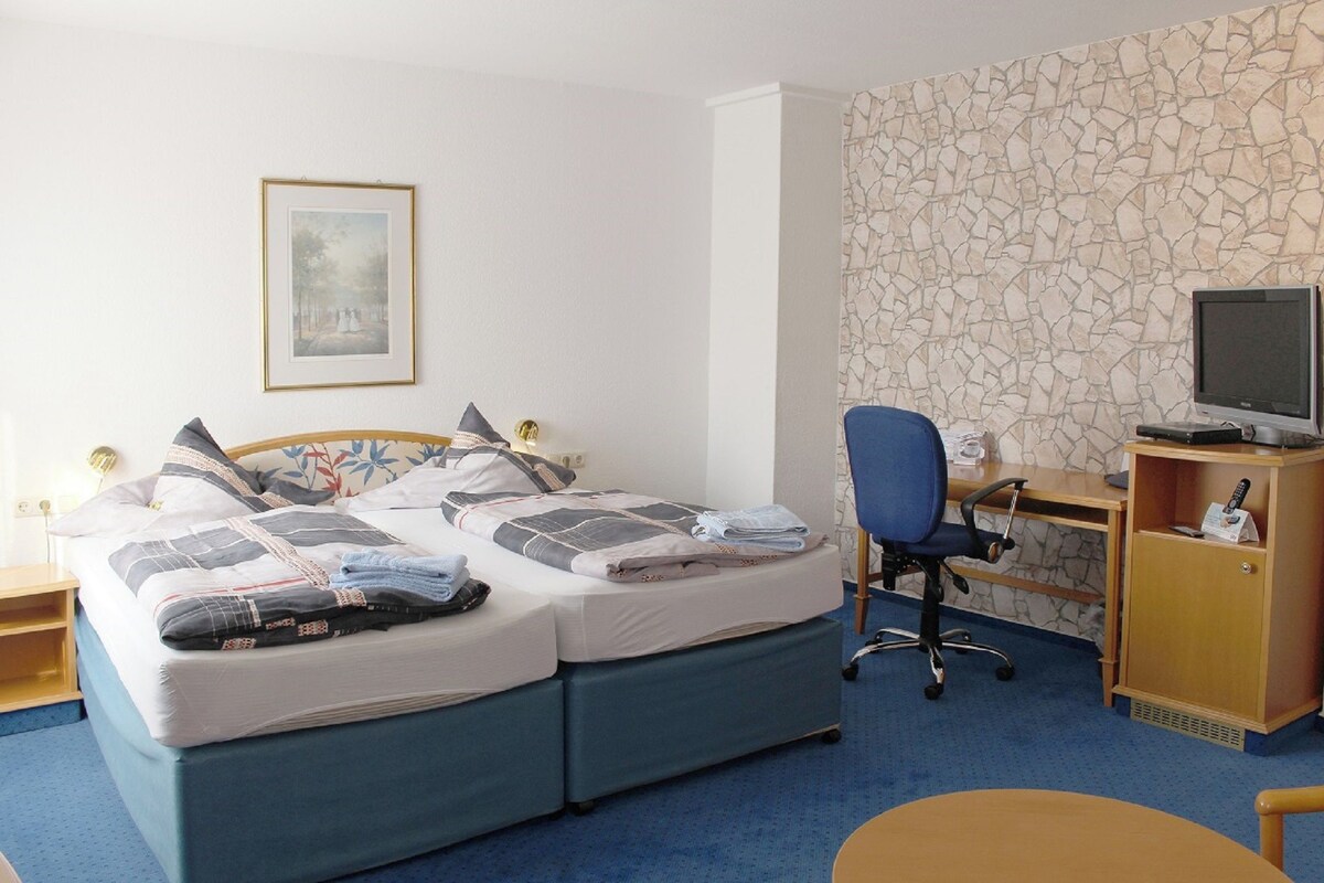 A hotel room with a double bed, a desk with a computer, a chair, and a TV on a stand.