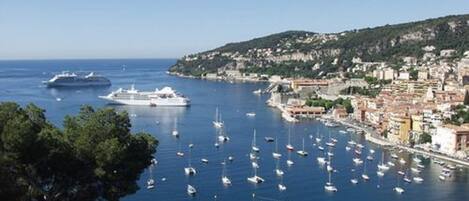 A coastal town with a marina filled with boats, a large cruise ship docked, and buildings along the waterfront.