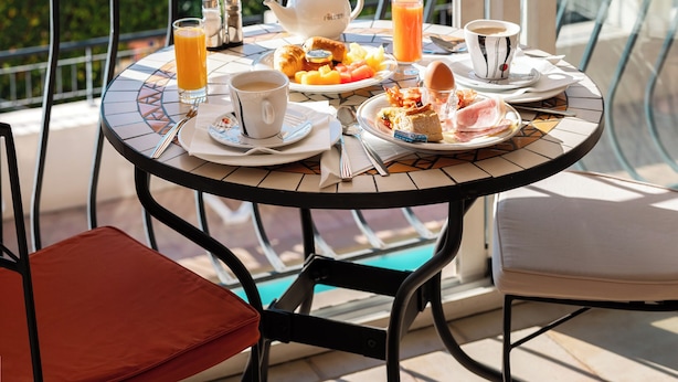 Une table ronde, dressée sur un balcon avec vue sur un plan d’eau et des bâtiments, est garnie d’aliments pour le petit-déjeuner, d’une théière et d’une tasse.
