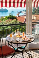 Una mesa redonda con alimentos para el desayuno, una tetera y una taza, ubicada en un balcón con vista a un cuerpo de agua y edificios.