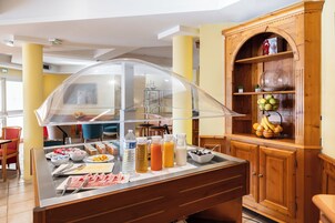 Zona de bufé con diversas opciones de comida y bebidas, un mueble de madera con frutas y un recinto de vidrio que cubre el bufé.