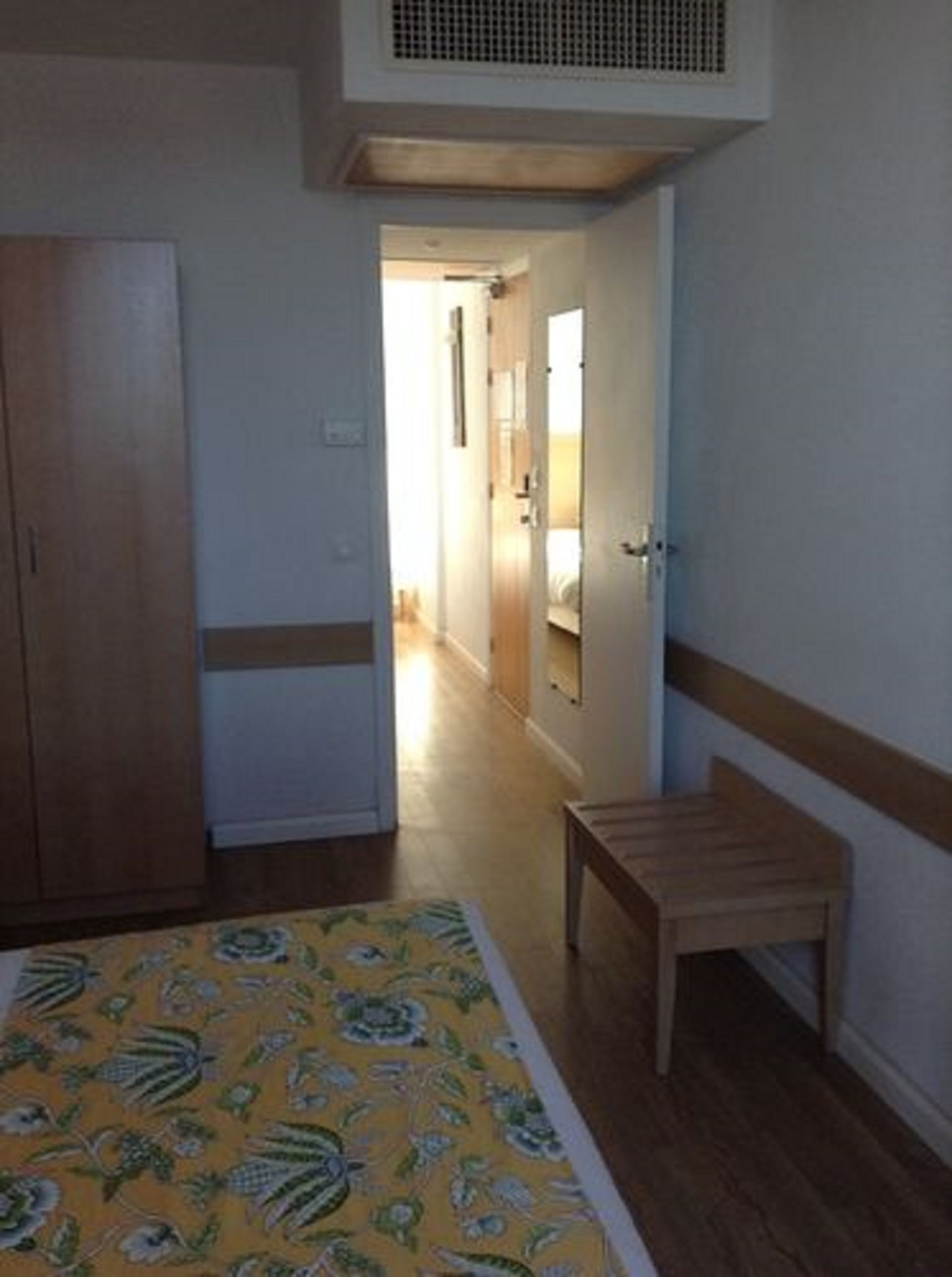 Quarto com cama, um banco de madeira, um guarda-roupa e um corredor que dá para outro espaço.