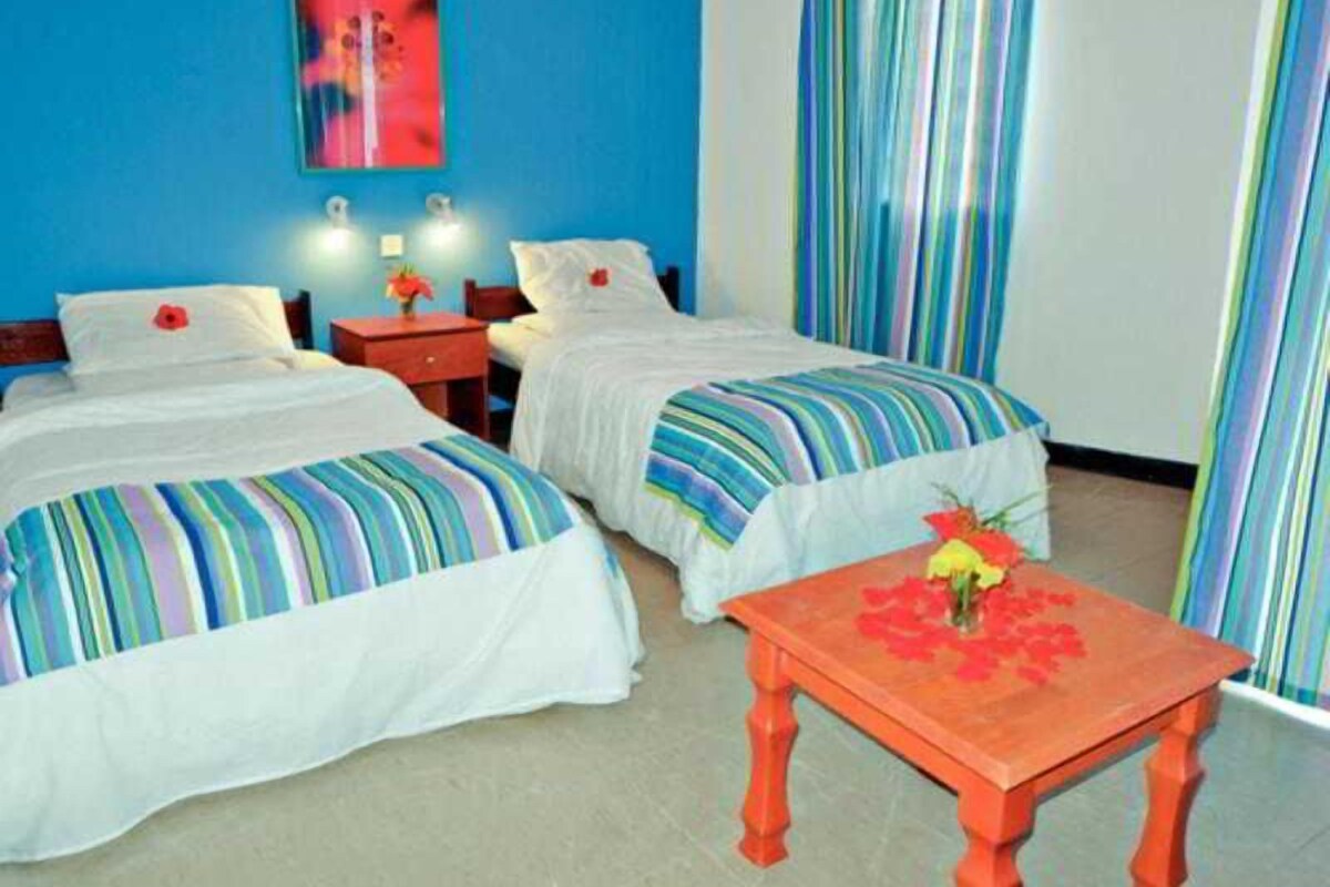 Habitación de hotel con dos camas, una mesita con un florero y una cortina a rayas coloridas.