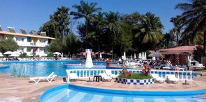 Zona de piscina con sillas de descanso, sombrillas y palmeras.