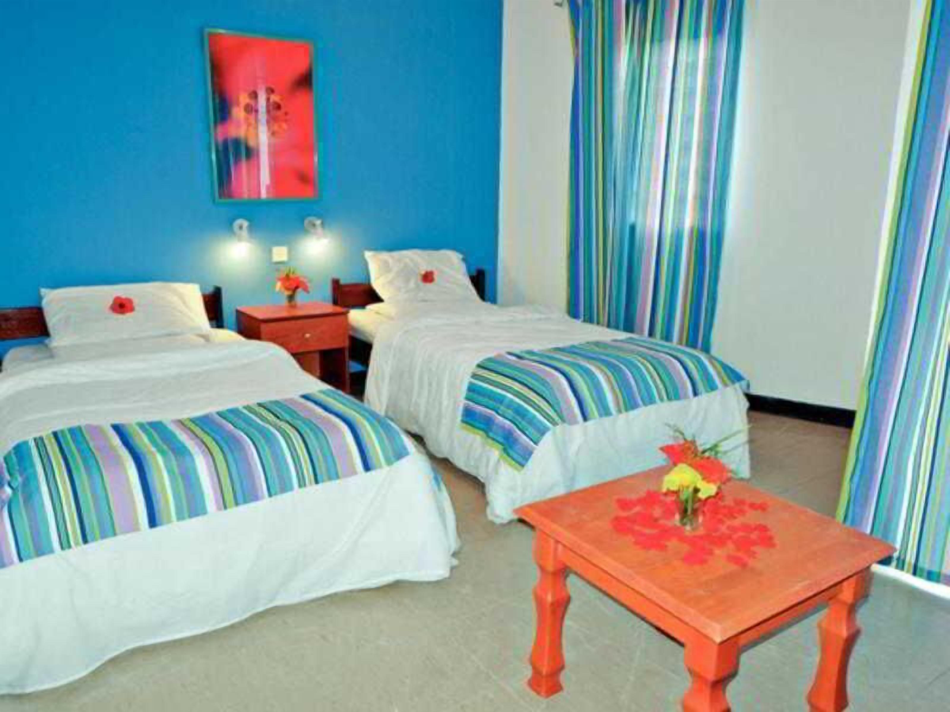 A hotel room with two beds, a small table with a vase of flowers, and a colorful striped curtain.