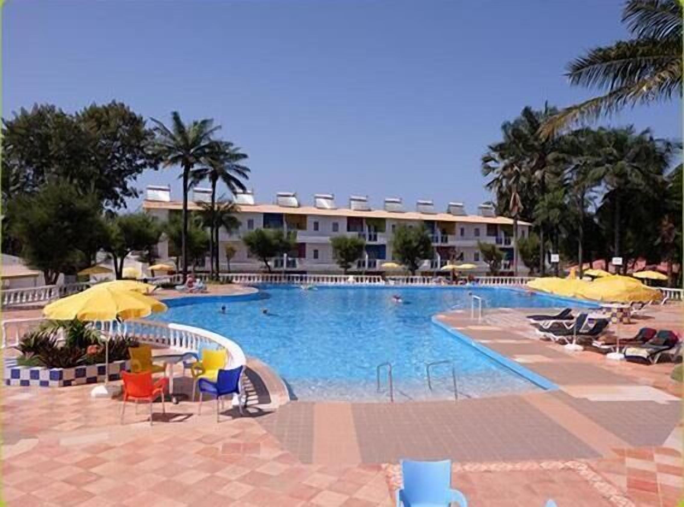 Una piscina con sillas de descanso y sombrillas, rodeada de palmeras y un edificio con varios balcones.