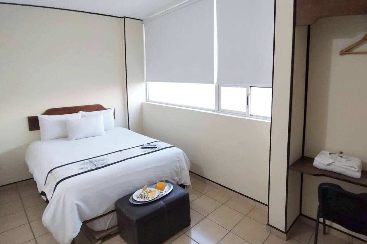 A hotel room with a bed, a small table with breakfast, a chair, a shelf with towels, and a window.