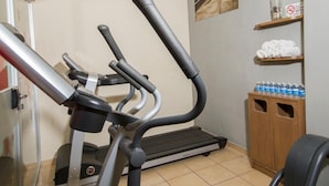 Une petite salle de sport équipée de deux tapis de course et d’un appareil elliptique. On y trouve également un meuble en bois contenant des serviettes et des bouteilles d’eau.