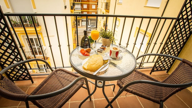Een balkon met een ronde tafel, twee rieten stoelen, een fles, een glas sinaasappelsap, een kleine potplant en een bord met eten.
