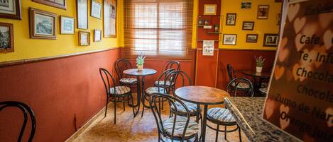 Un café avec de nombreuses tables et chaises, des tableaux encadrés aux murs et une fenêtre avec des stores.