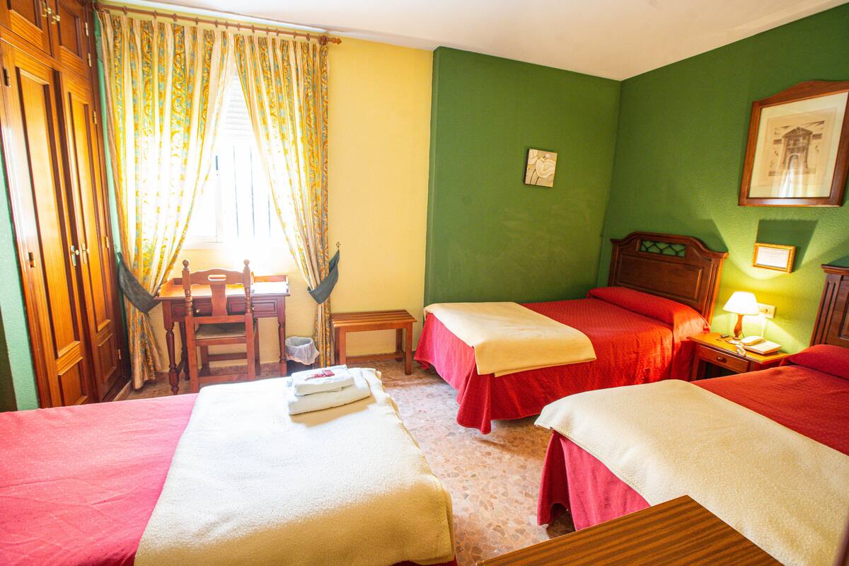 Quarto de hotel com duas camas, uma escrivaninha, uma cadeira e uma mesinha.