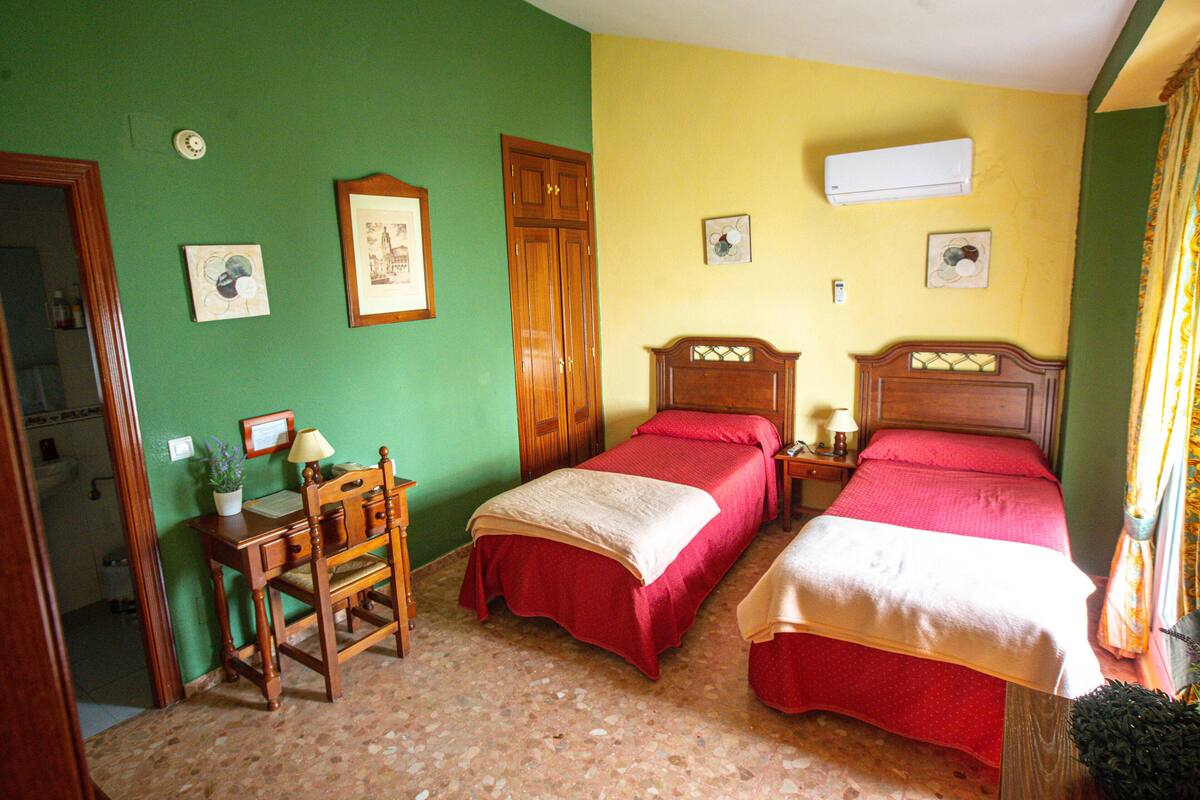 Quarto com duas camas, uma escrivaninha com cadeira, guarda-roupa de madeira e ar-condicionado fixado na parede.