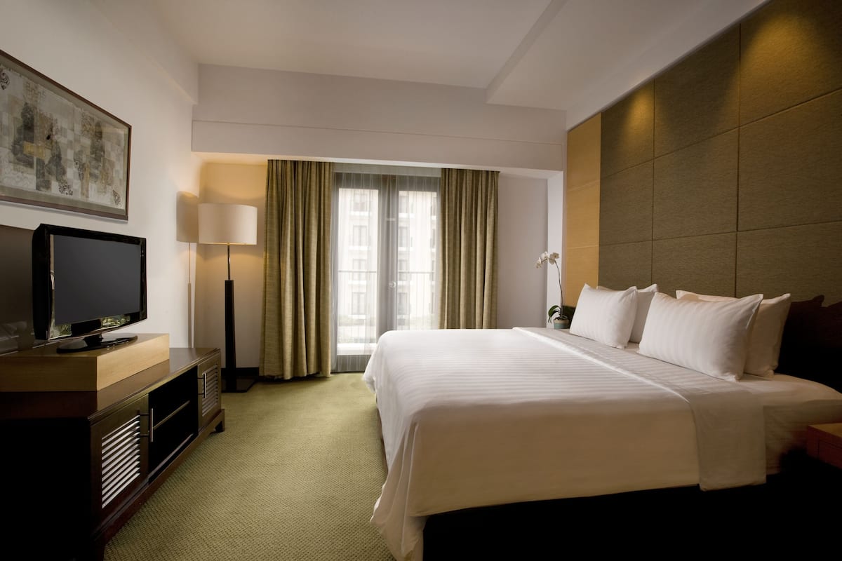 Ein Hotelzimmer mit einem großen Bett, einem Fernseher auf einer Holzkonsole, einer Stehlampe und einem Fenster mit Vorhängen.