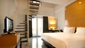 A hotel room with a large bed, a TV on a wooden stand, a desk with a lamp, a mirror, and a staircase leading to another floor.