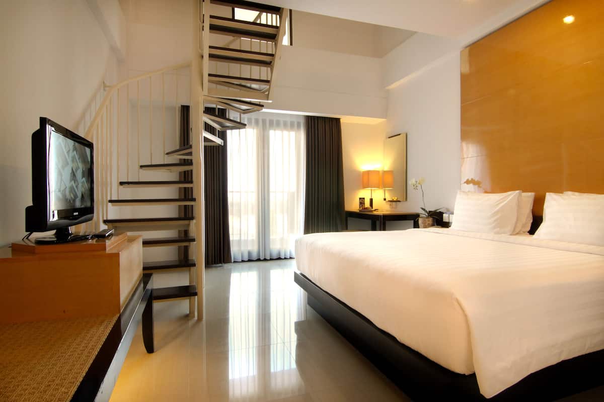 Ein Hotelzimmer mit einem großen Bett, einem Fernseher auf einem Holzbalken, einem Schreibtisch mit Lampe, einem Spiegel und einer Treppe, die zu einer anderen Etage führt.
